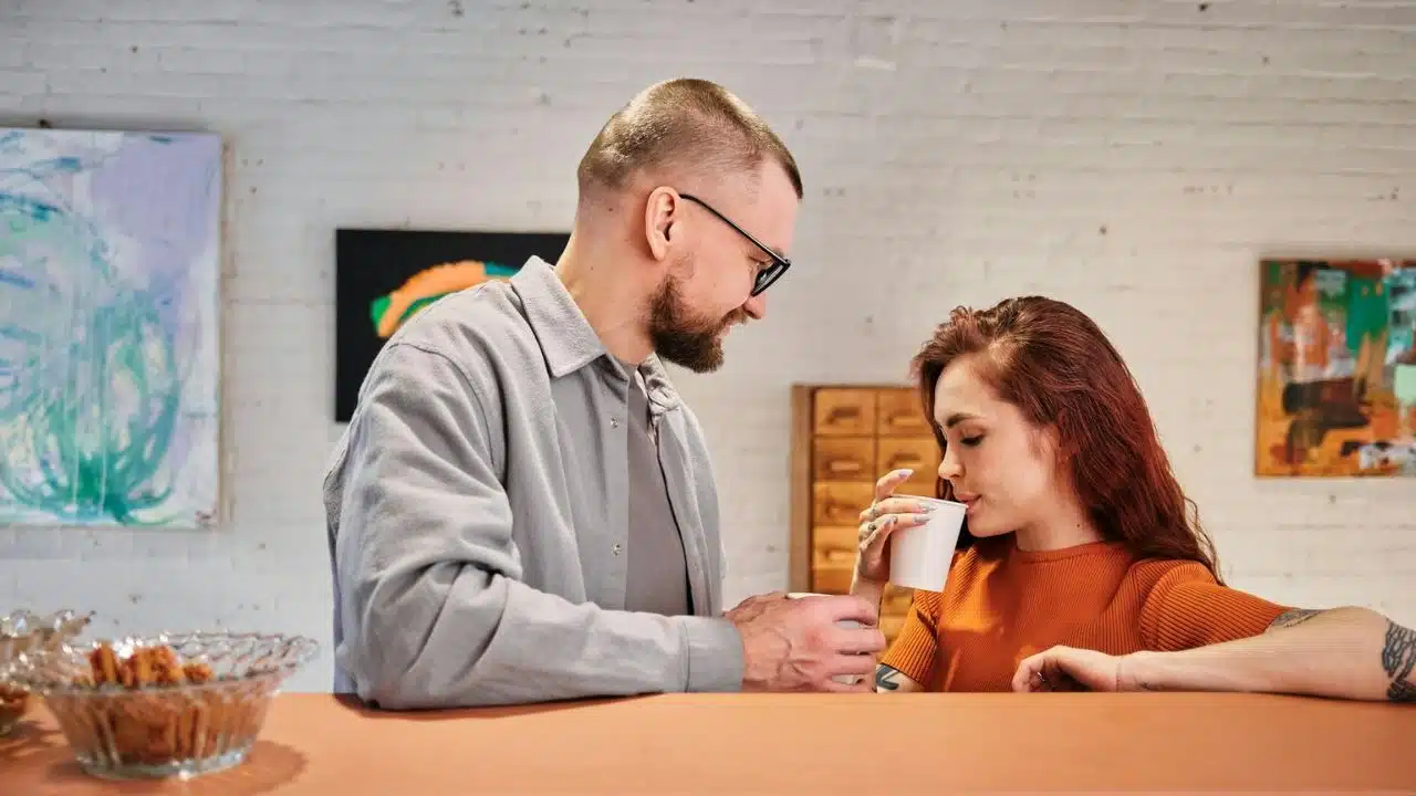 A man with a beard and glasses looks at a woman with red hair, who sips from a cup.