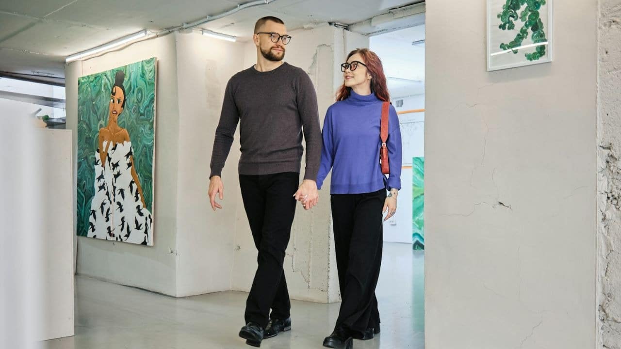 A man and woman, holding hands, walk through an art gallery, looking at each other.