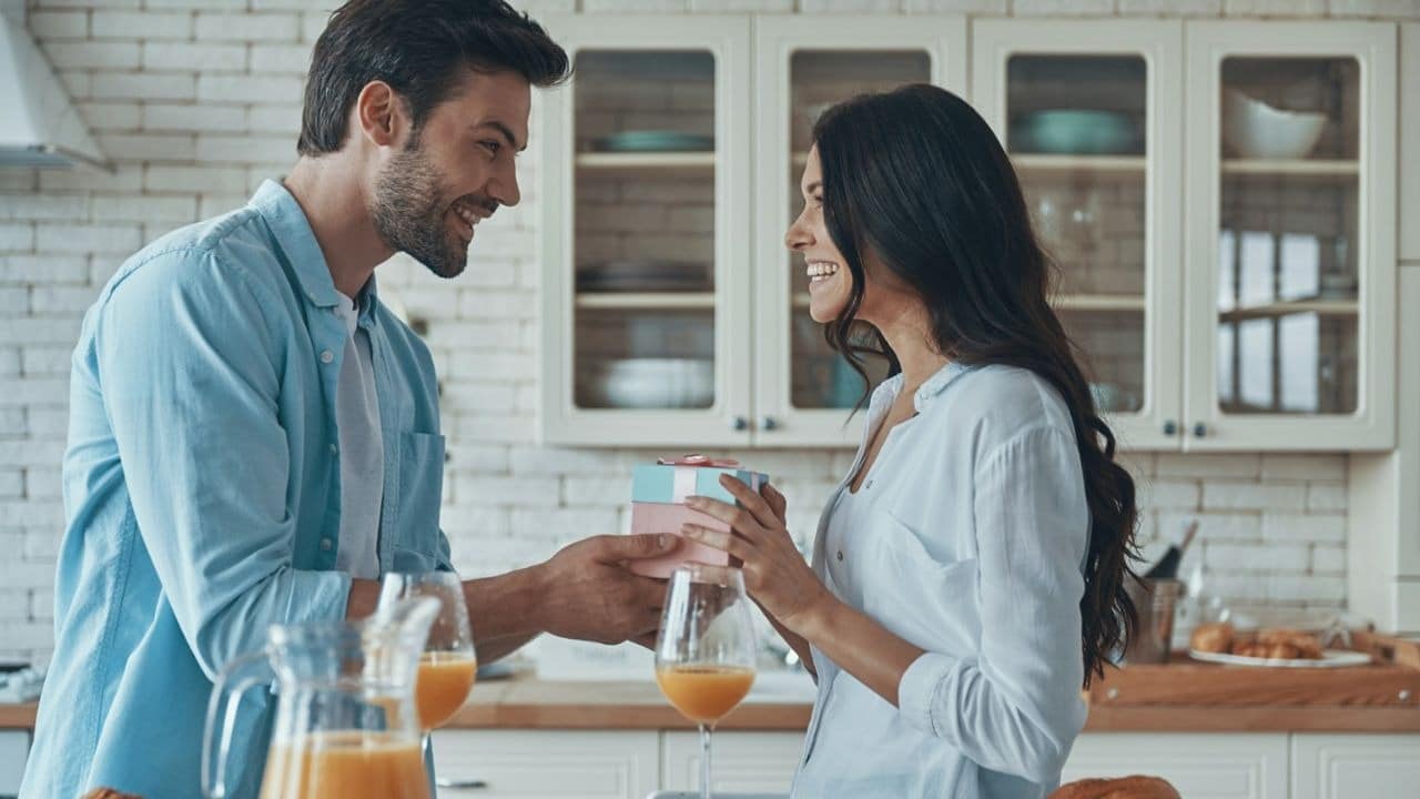 A smiling man gives a wrapped gift to a smiling woman in a kitchen setting.