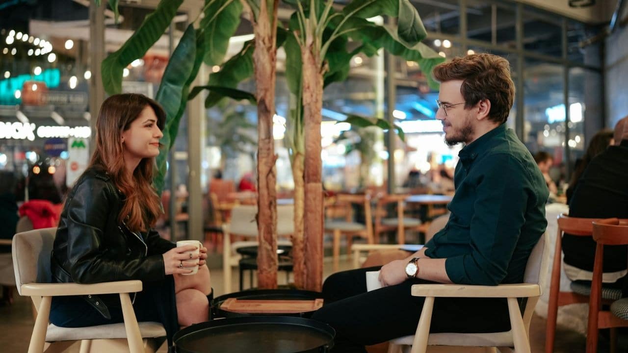 A man and woman sit facing each other in a cafe, talking and holding drinks.