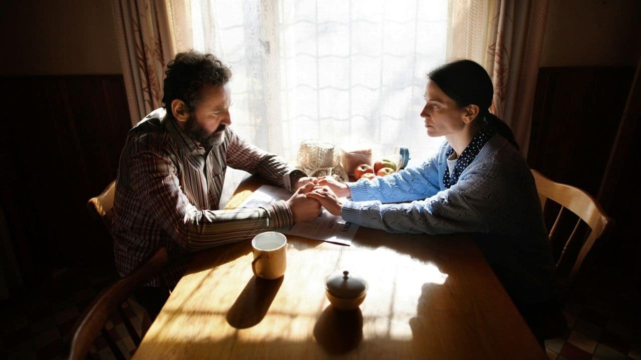 A man and woman hold hands across a table, silhouetted against a bright window.