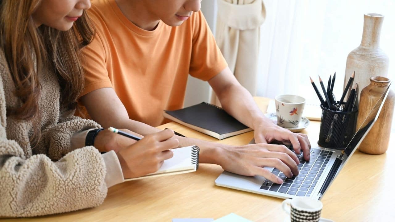 A man types on a laptop while a woman writes in a notebook beside him.