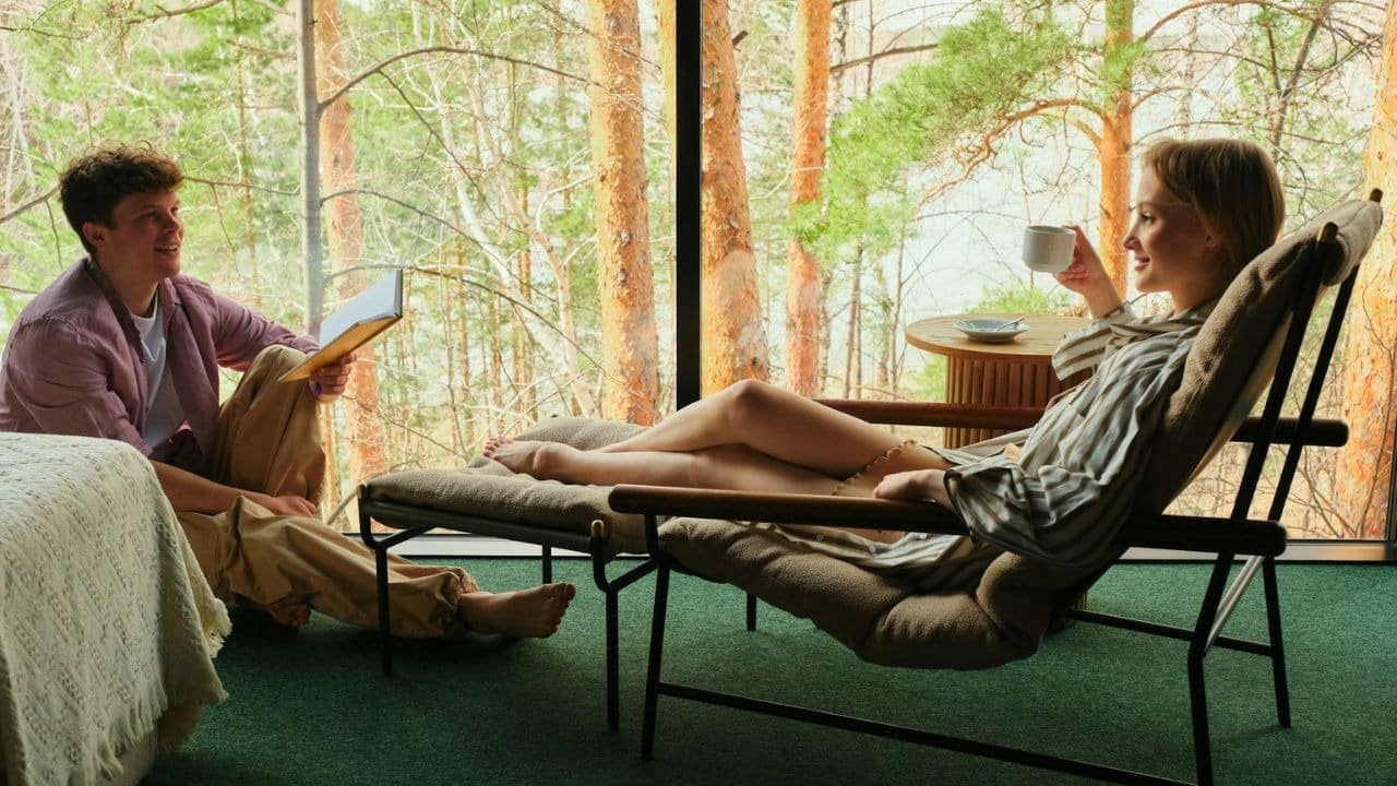 A couple relaxes indoors with coffee and a book, enjoying the forest view outside.