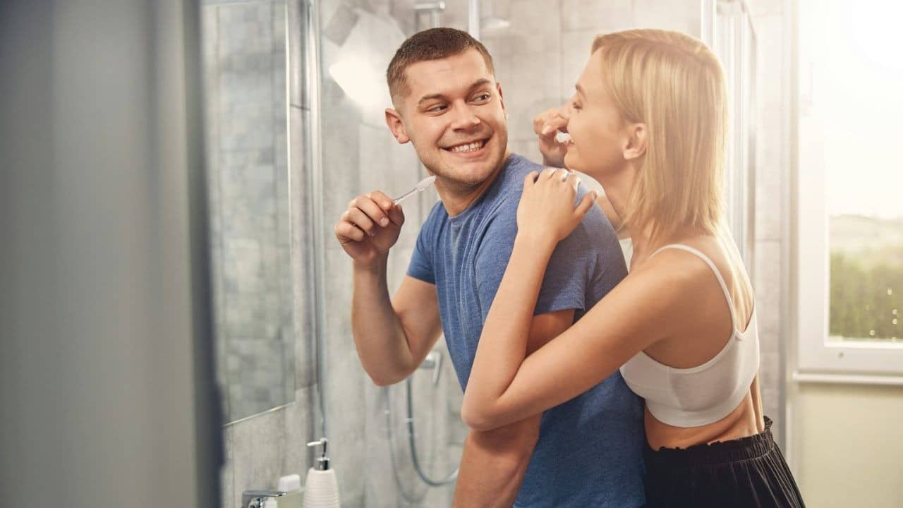 A smiling couple brushes their teeth together in a bathroom, looking at each other.