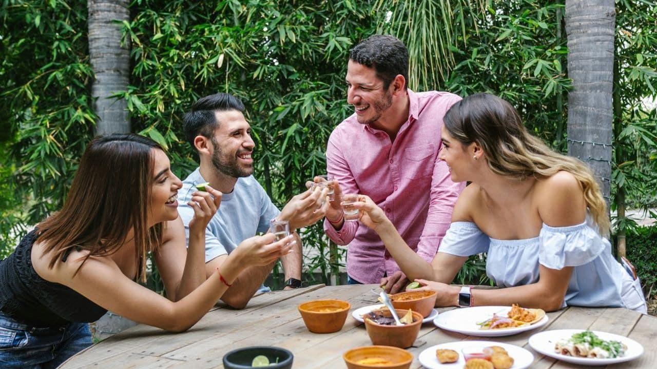 Four smiling friends raise shot glasses at a wooden table outdoors with food.