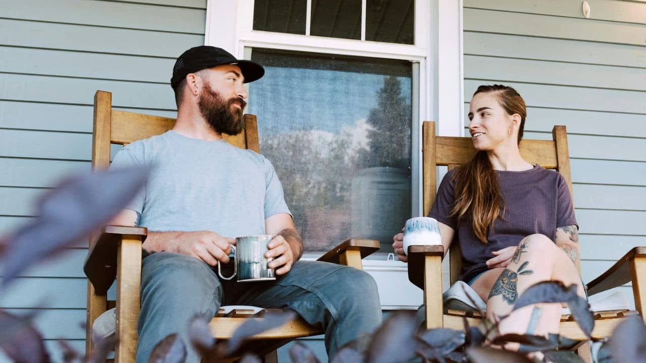 A man and a woman sit in rocking chairs on a porch, holding mugs and conversing.