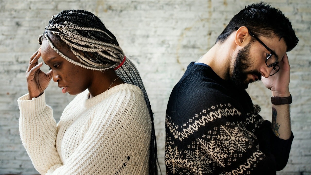 A couple standing with the backs to each other and holding their hands up to their foreheads.