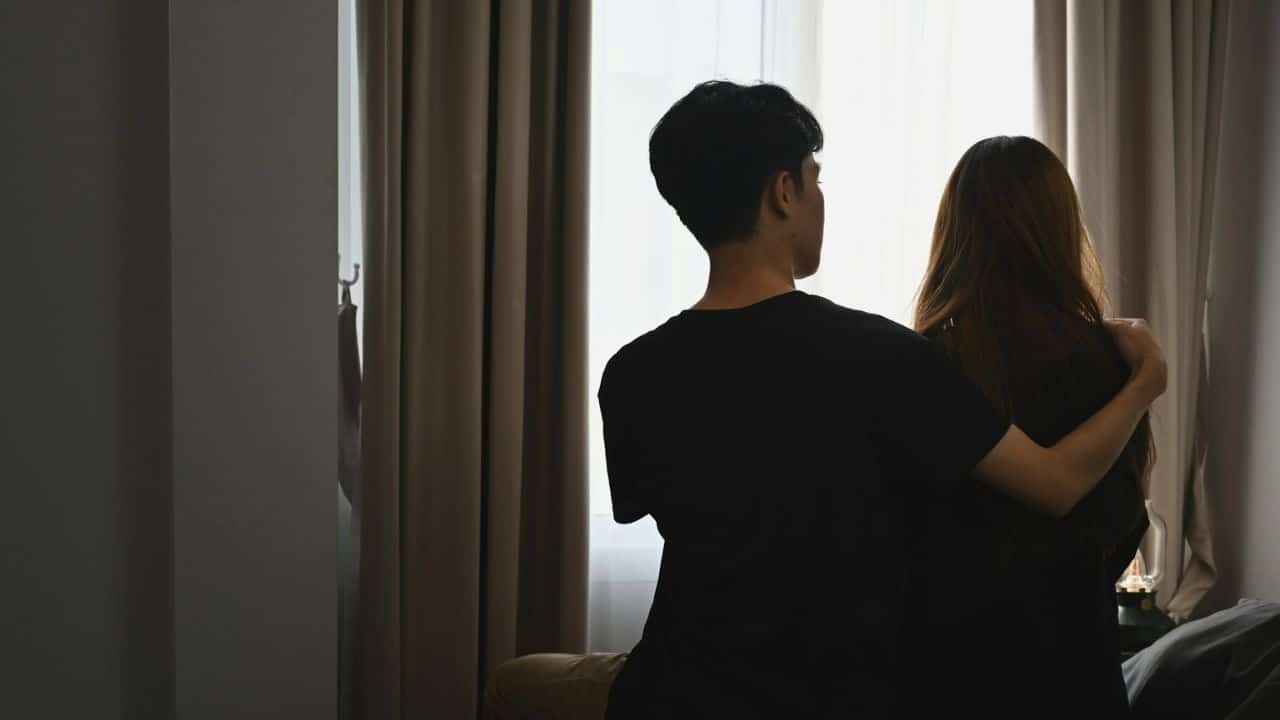 A person with their arm around another person's shoulder looks out a window.