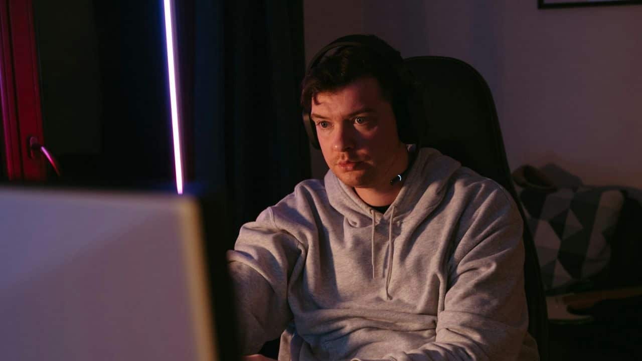 A young man with headphones on looks intently at a computer screen in a dimly lit room.