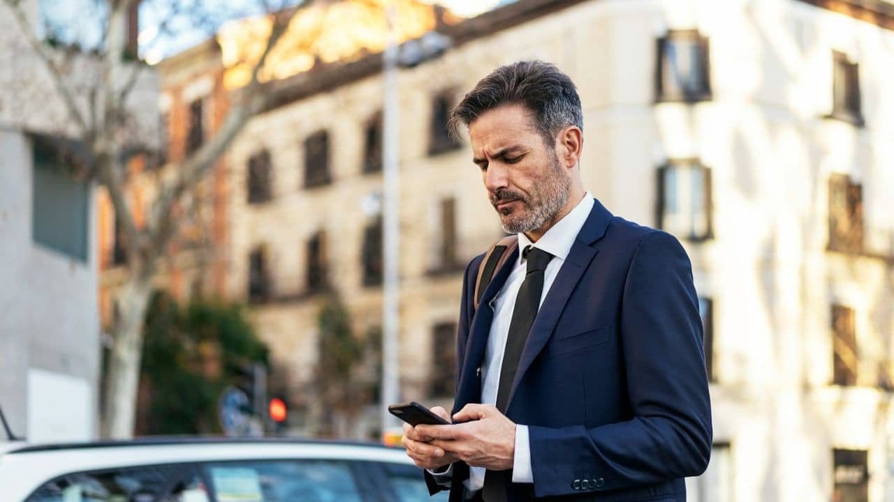 A man in a suit looks down at his phone while standing on a city street.