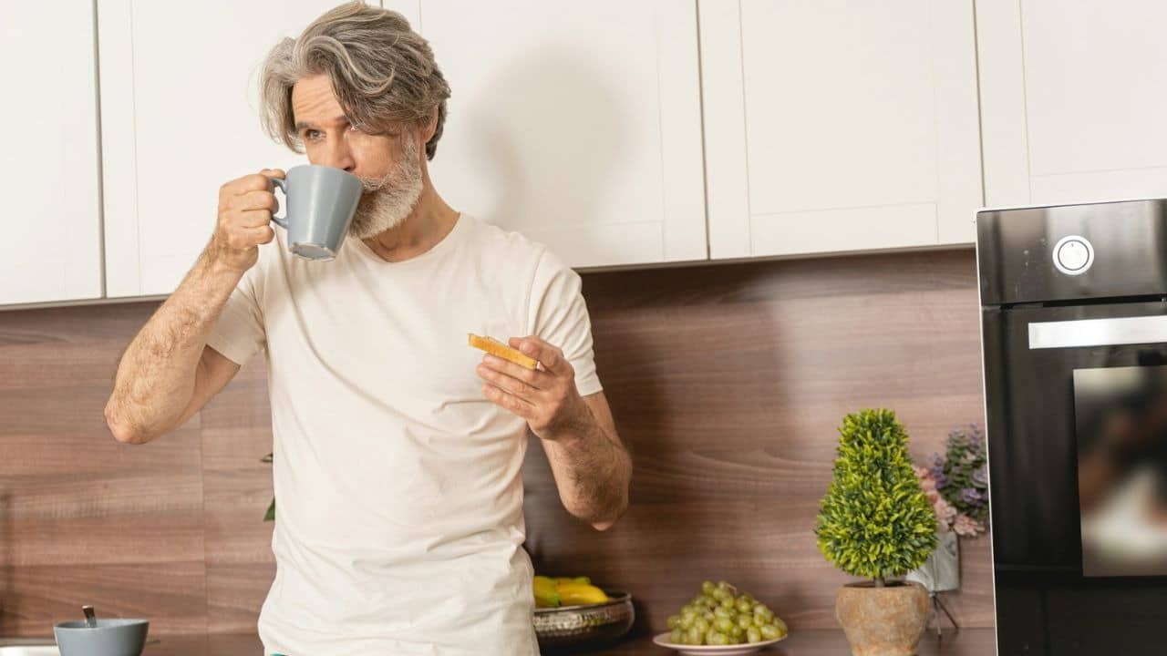 A grey-haired man with a beard sips from a mug, holding food in the other hand.