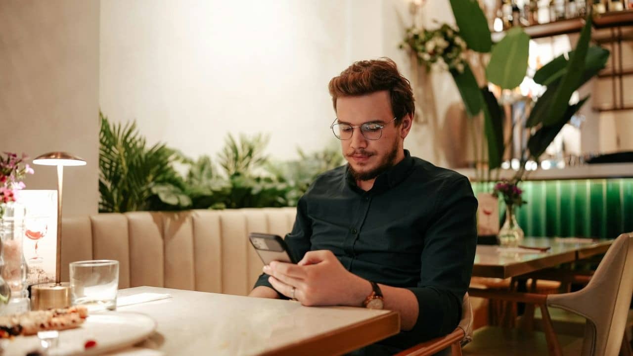 A man with glasses and a beard sits at a restaurant table, looking at his phone.