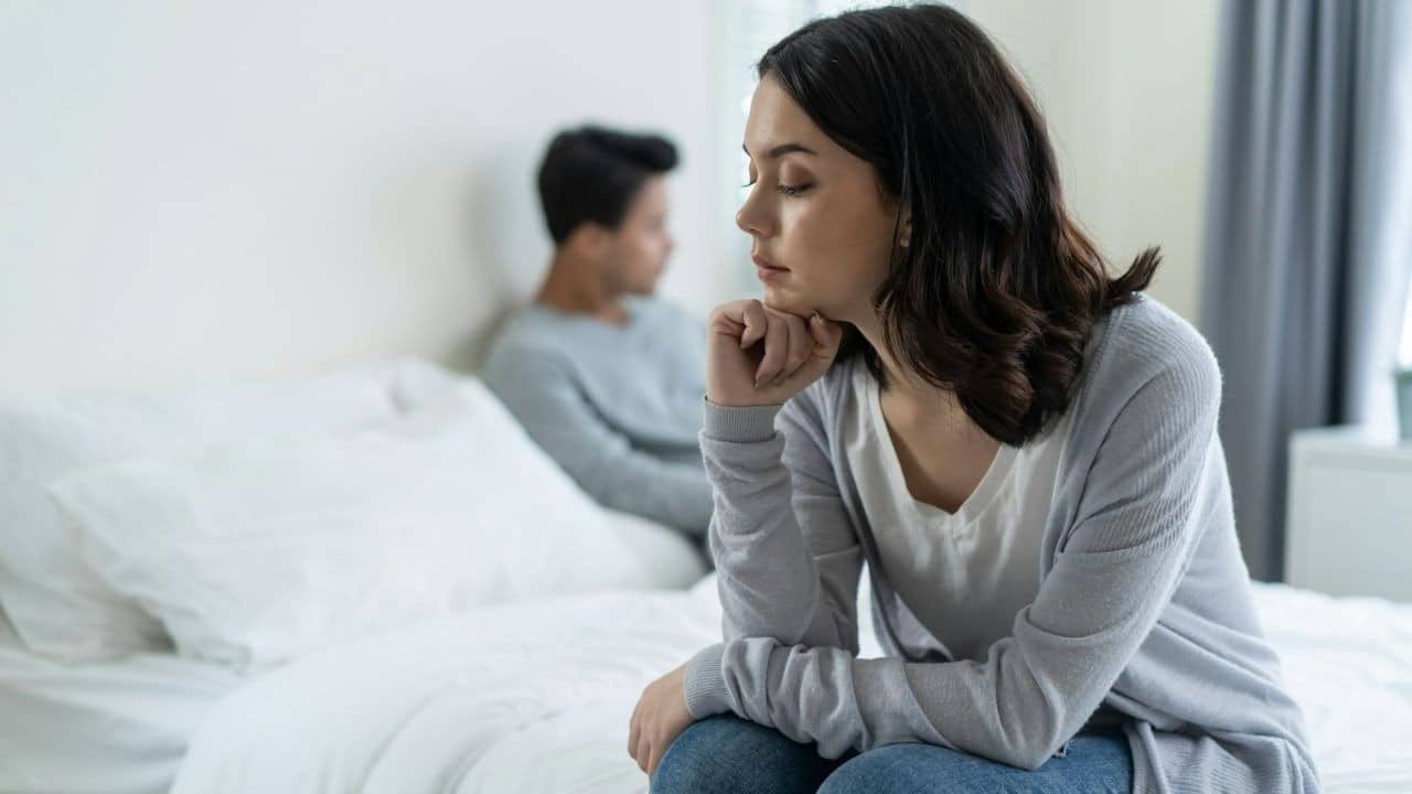 A thoughtful woman sits on a bed, while a man looks away in the background.