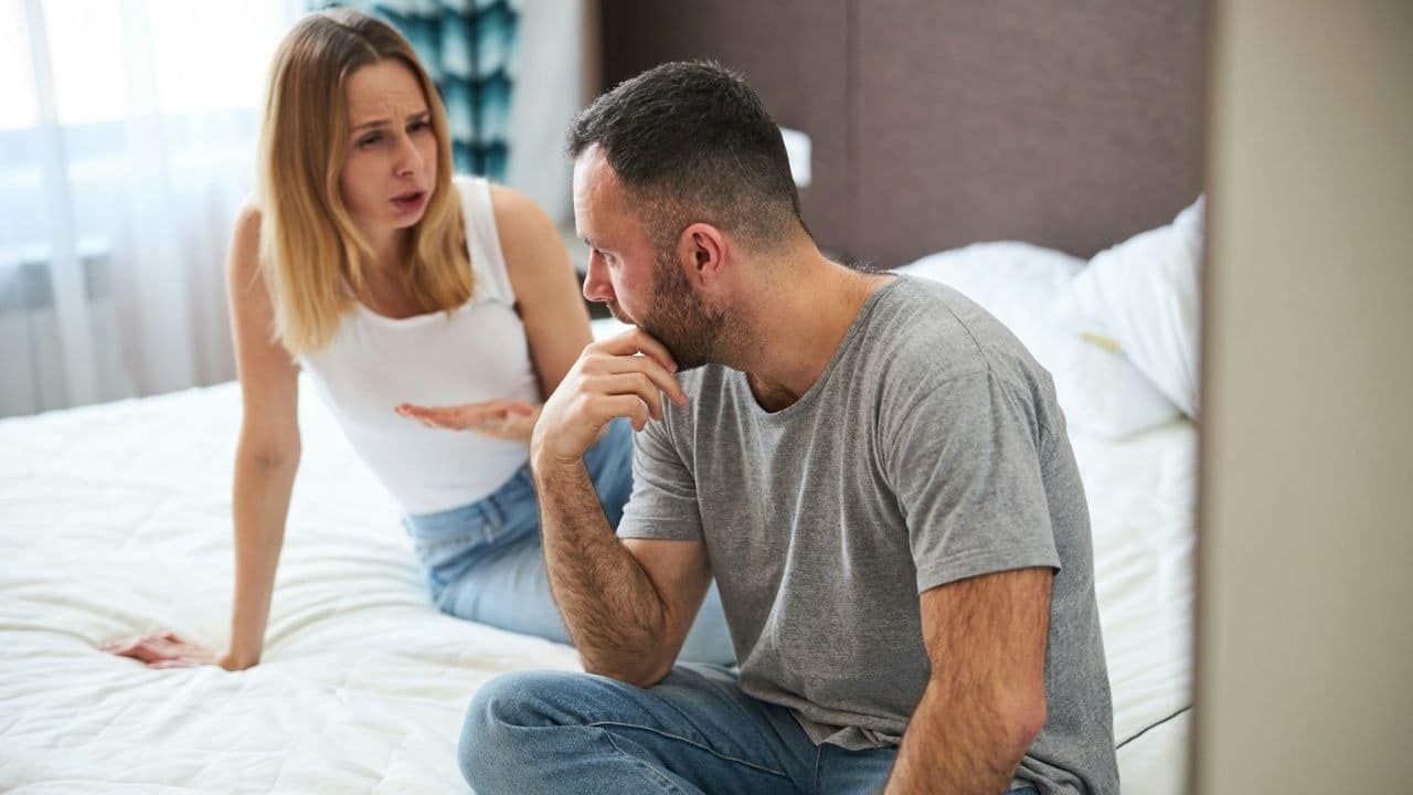 A man sits on a bed with his hand on his chin, while a woman in the background gestures and talks.