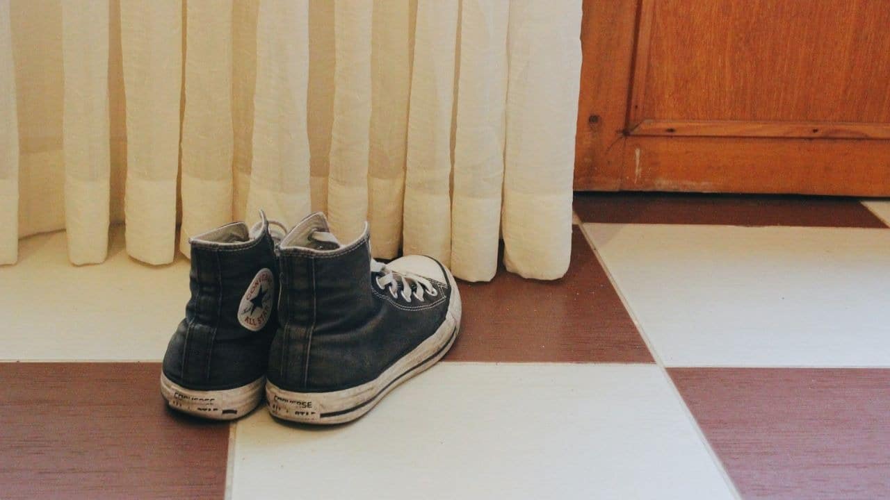 A pair of black Converse high-top sneakers sits on a tiled floor near a white curtain and wooden door.