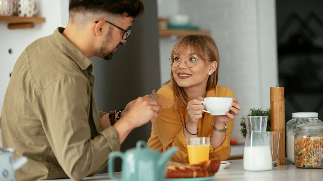 A man spreads something on food while a woman holding a cup smiles at him.