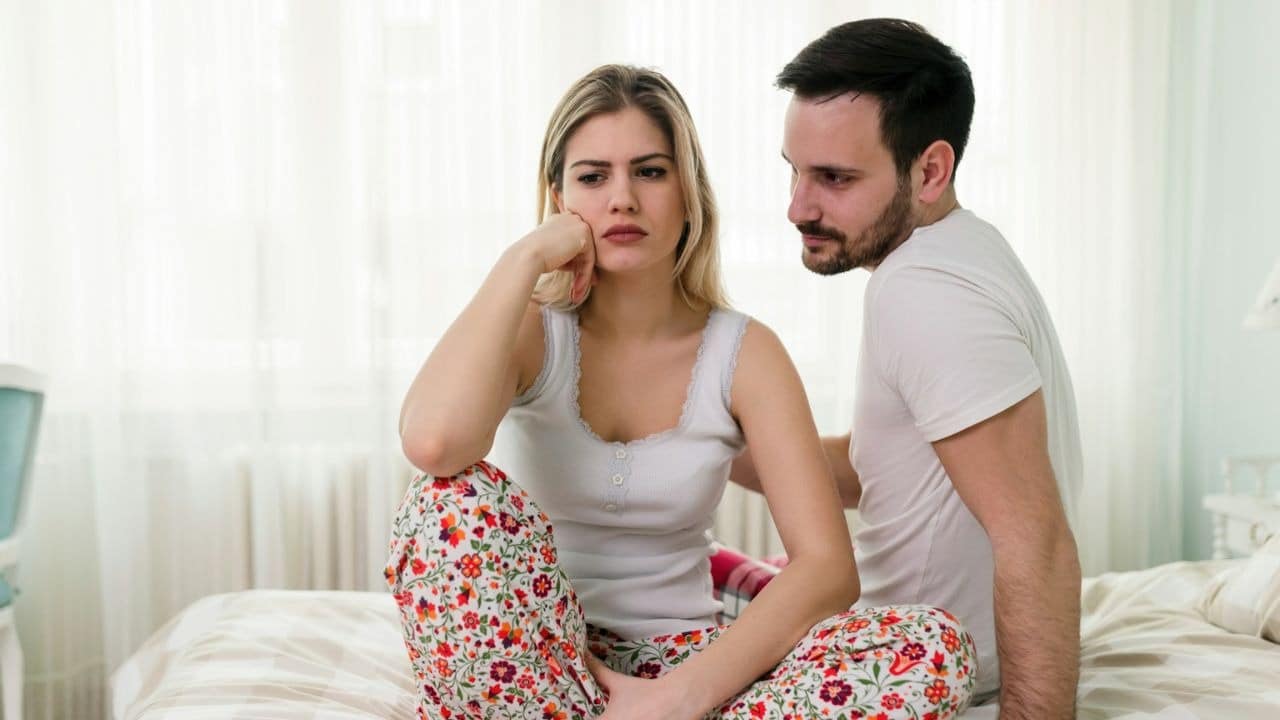 A man looks concernedly at a woman with a sad expression, who is sitting on a bed.