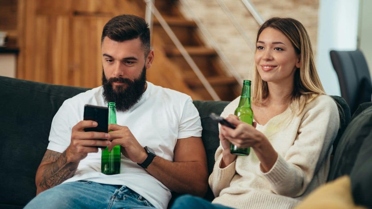 A man looks at his phone while a woman watches TV, both holding green bottles.