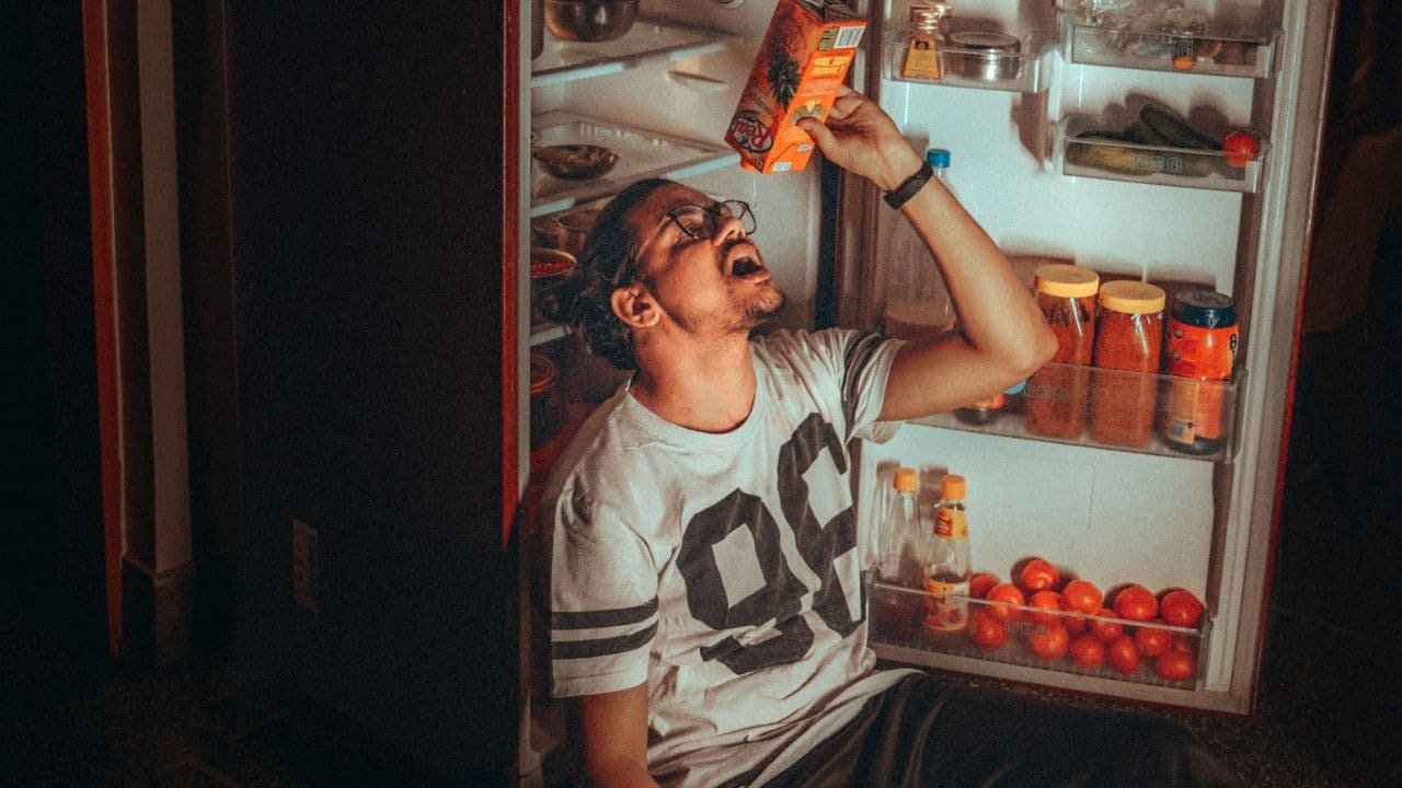 A man sits on the floor with an open refrigerator, drinking from a carton.