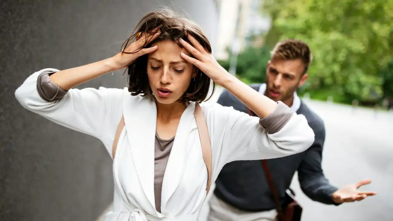 A woman holds her head with both hands while a man gestures and speaks behind her.