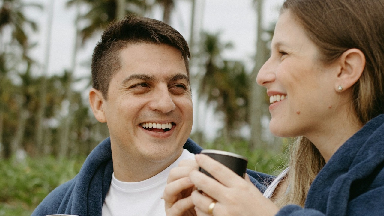 A man is smiling at a woman who’s having coffee and smiling.