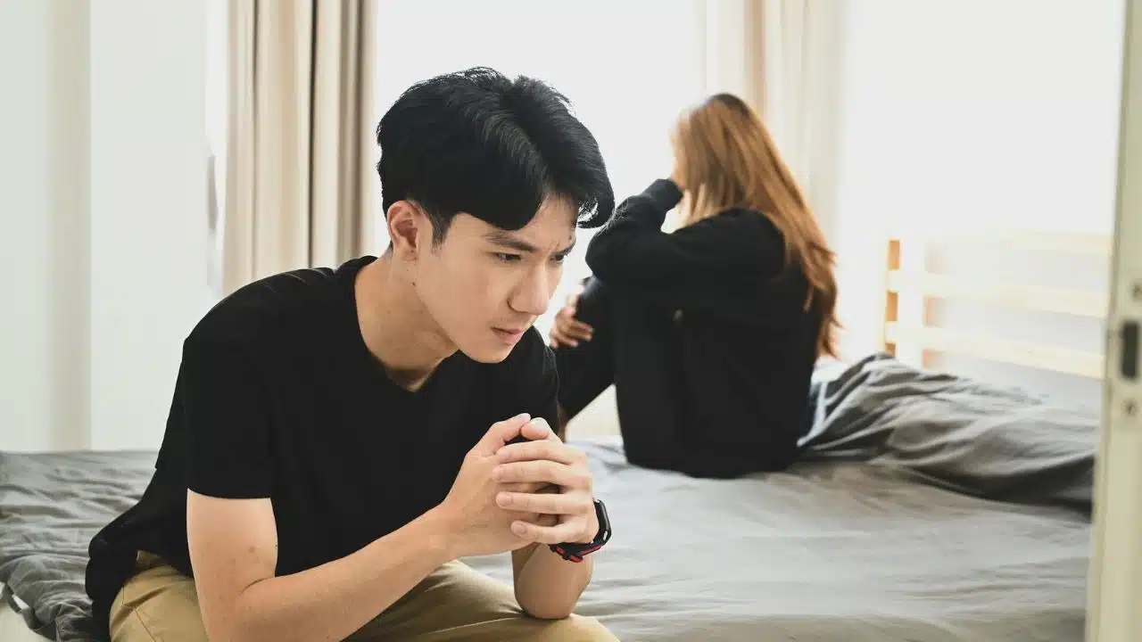 A pensive man sits in the foreground with a sad, crying woman in the background.