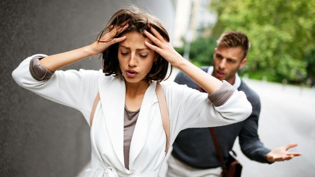 A stressed woman holds her head as a frustrated man gestures behind her on a street.