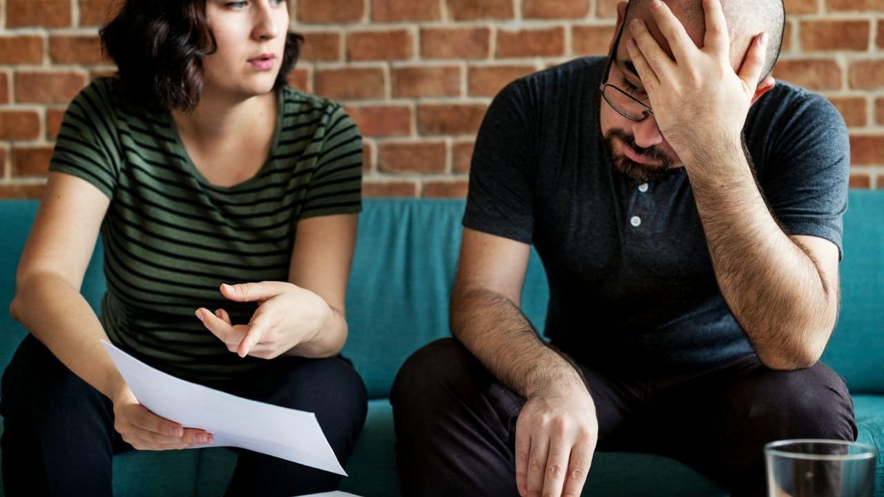 A stressed man is sitting on a couch holding his head, next to a woman holding papers and talking.