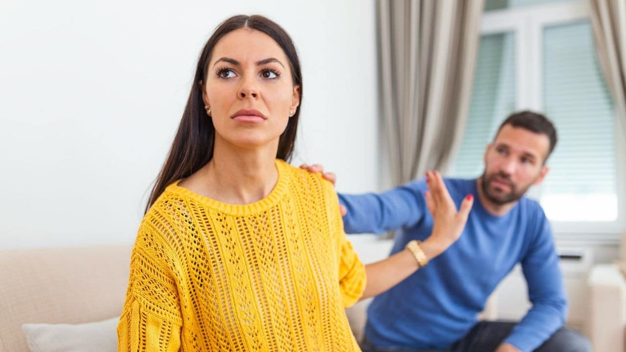 A woman in a yellow sweater pushes away a man in blue with an upset expression
