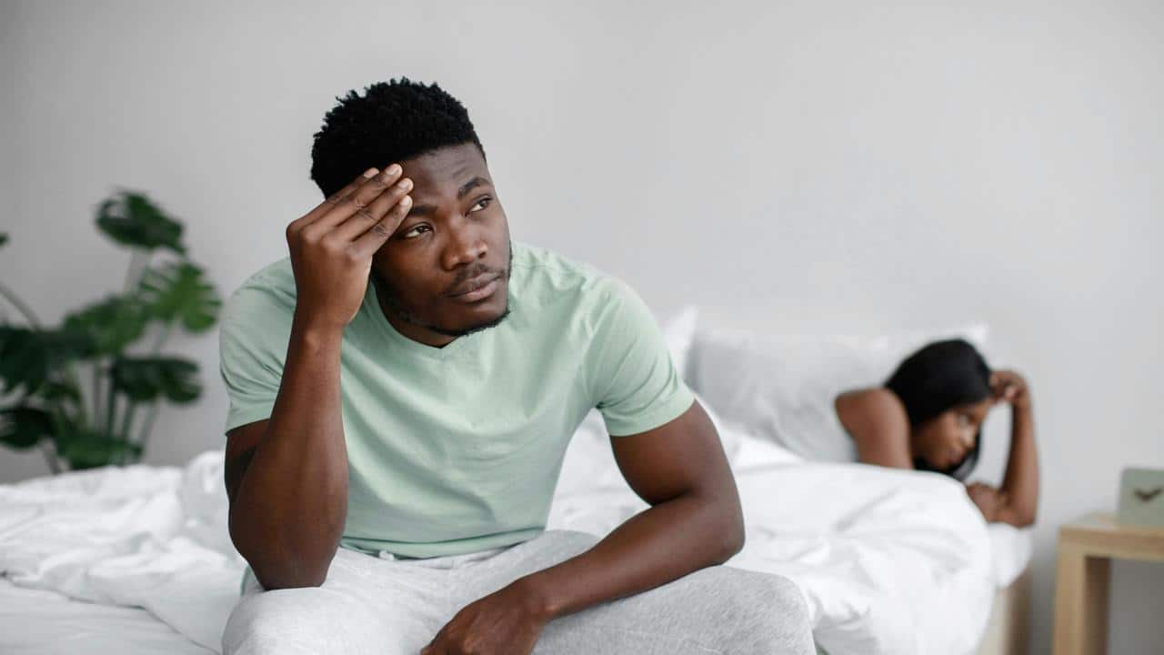 A man sitting on a bed looking stressed while a woman lies behind him appearing upset.