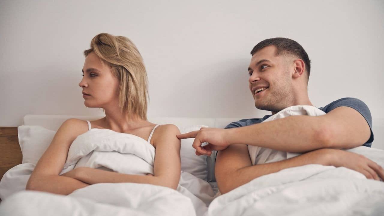 A man in bed is smiling and poking a woman who has her arms crossed and is looking away.