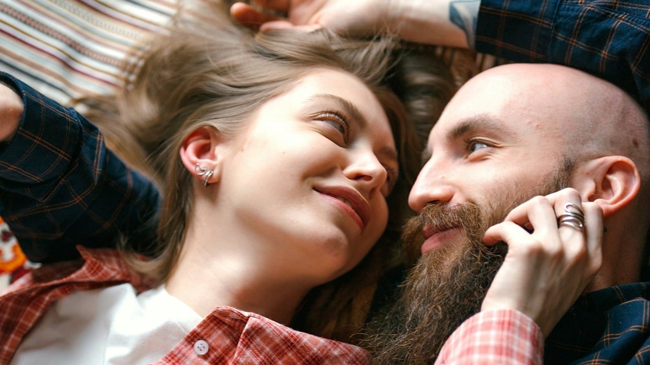 A bald man lying on the floor with a woman who’s stroking his beard.