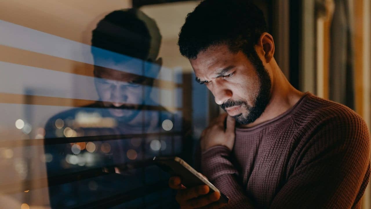 A concerned man with a beard stands by a window at night, looking at his phone.