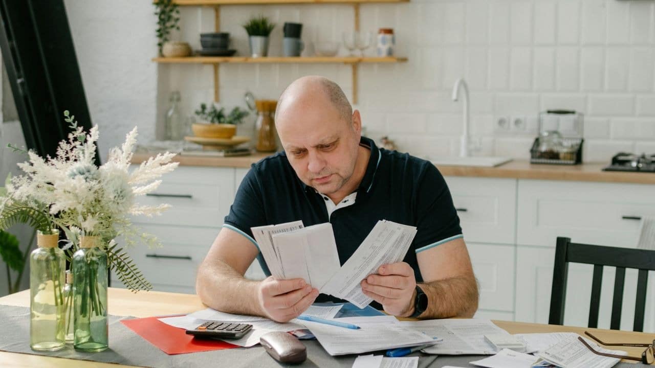 A serious bald man sits at a table, looking at papers and a calculator.