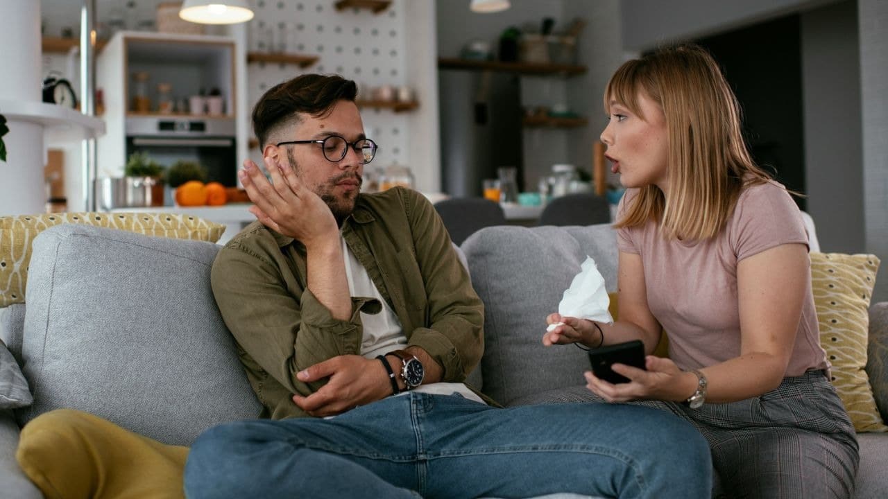 A woman talks animatedly to a man who is sitting on a couch with a bored expression.