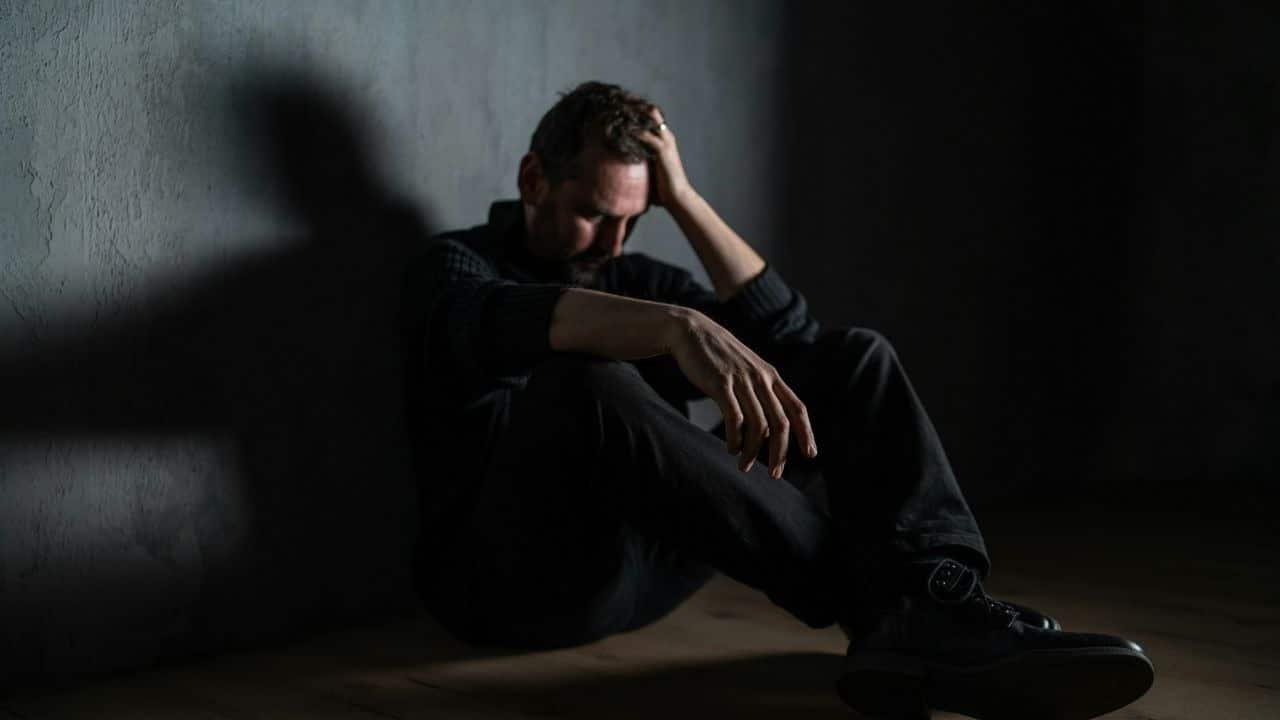 A stressed man sits against a wall, holding his head with his hand and looking down.