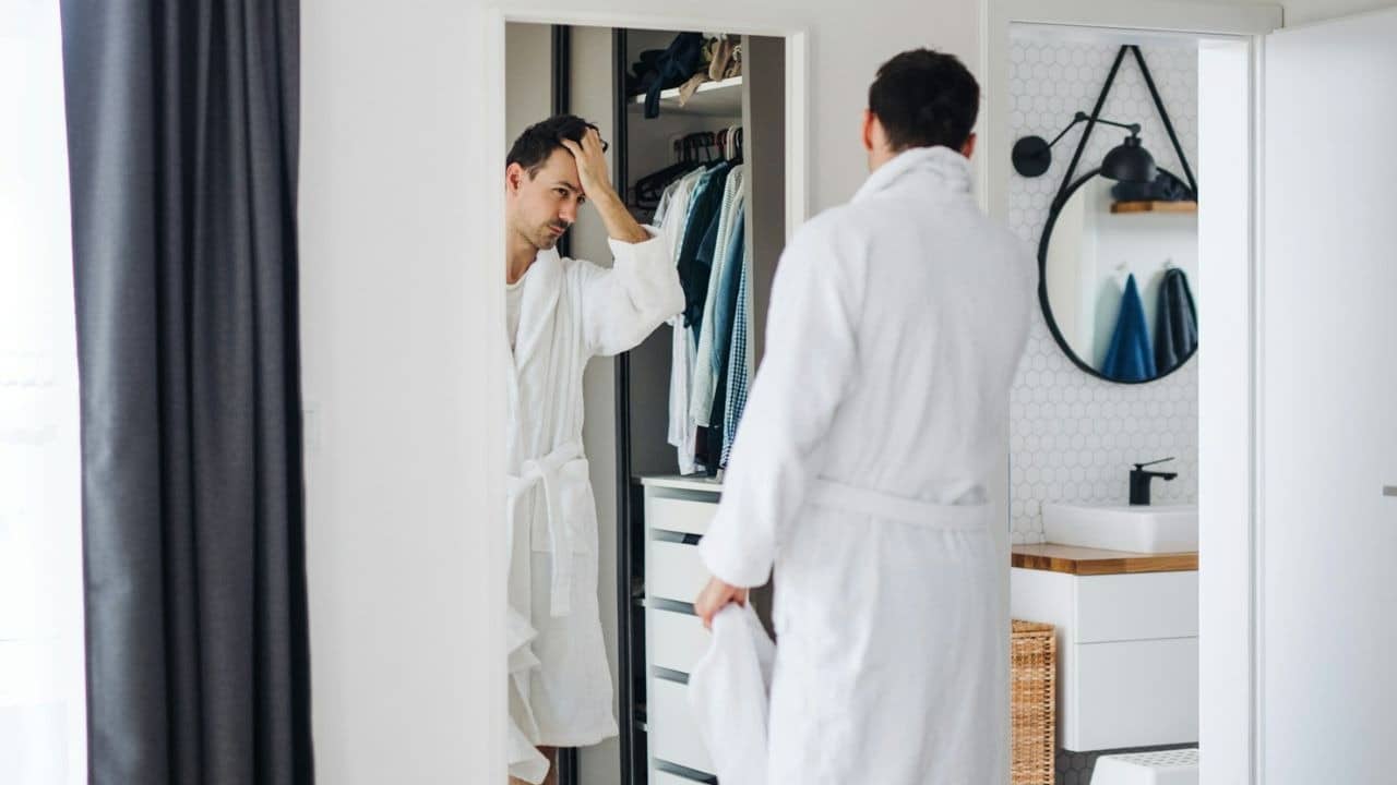A man in a white robe looks in a mirror, adjusting his hair in a closet.