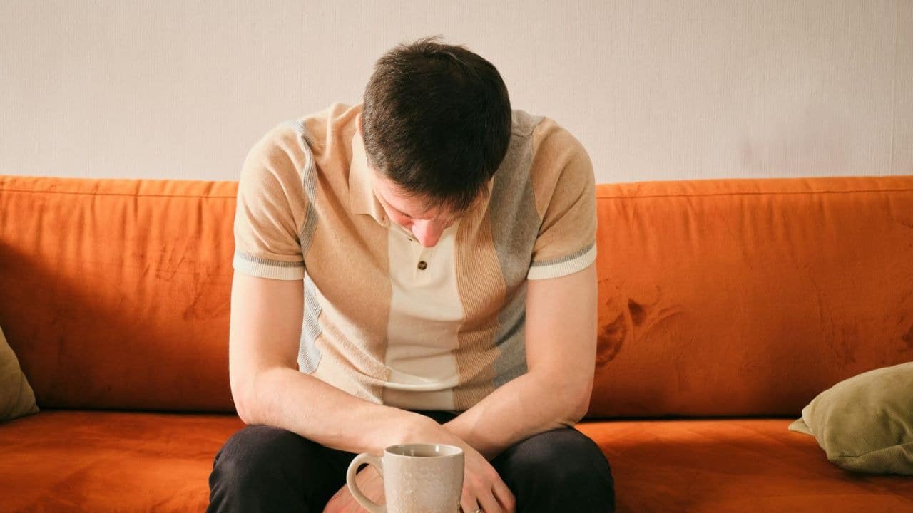 A man sits on an orange couch, looking down at his lap with a mug in front of him.