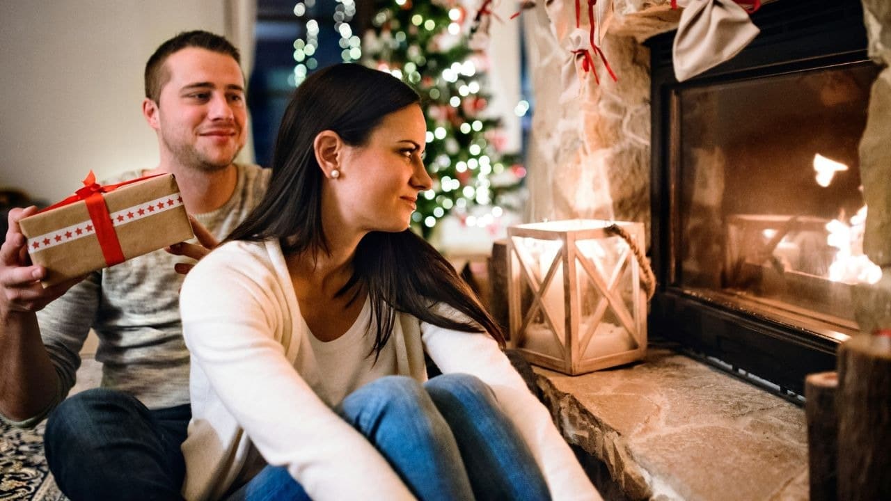 A couple sits on the floor near a fireplace and a Christmas tree, with the man holding a wrapped gift.
