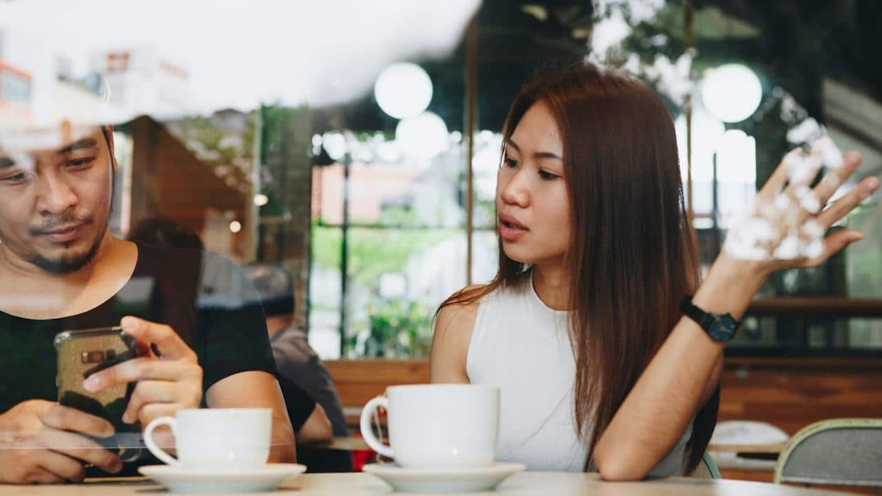 A man looking at his phone while a woman sitting across from him at a cafe table is talking and gesturing with her hand.