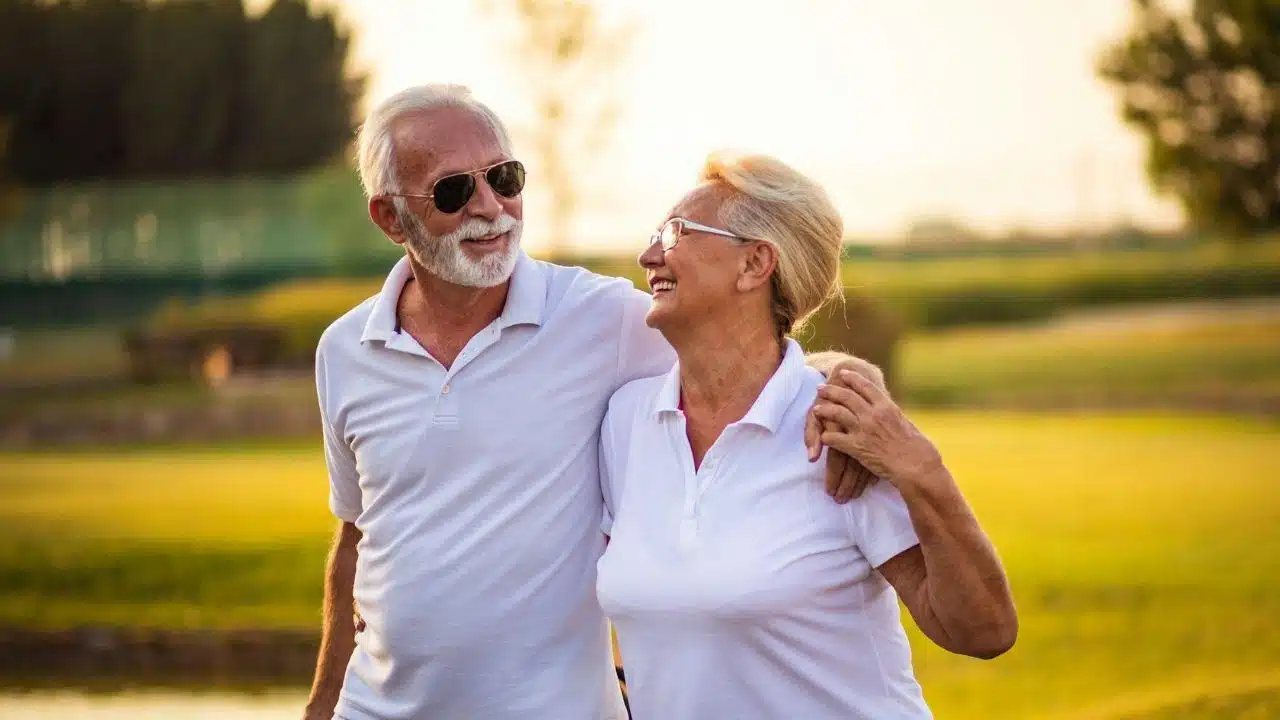 A cheerful elderly couple, arm-in-arm, smiles at each other outdoors.
