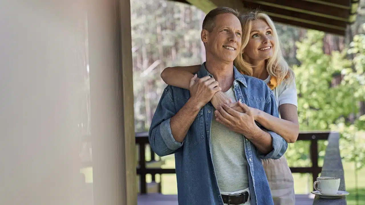 A smiling woman embracing a man while they are enjoying the morning with hot drinks on the terrace of a holiday home.