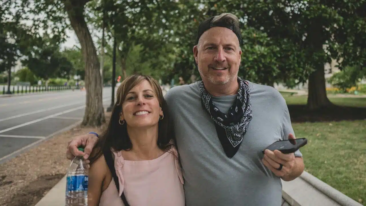 A cheerful couple smiling directly at the camera while walking side-by-side along a tree-lined city sidewalk.