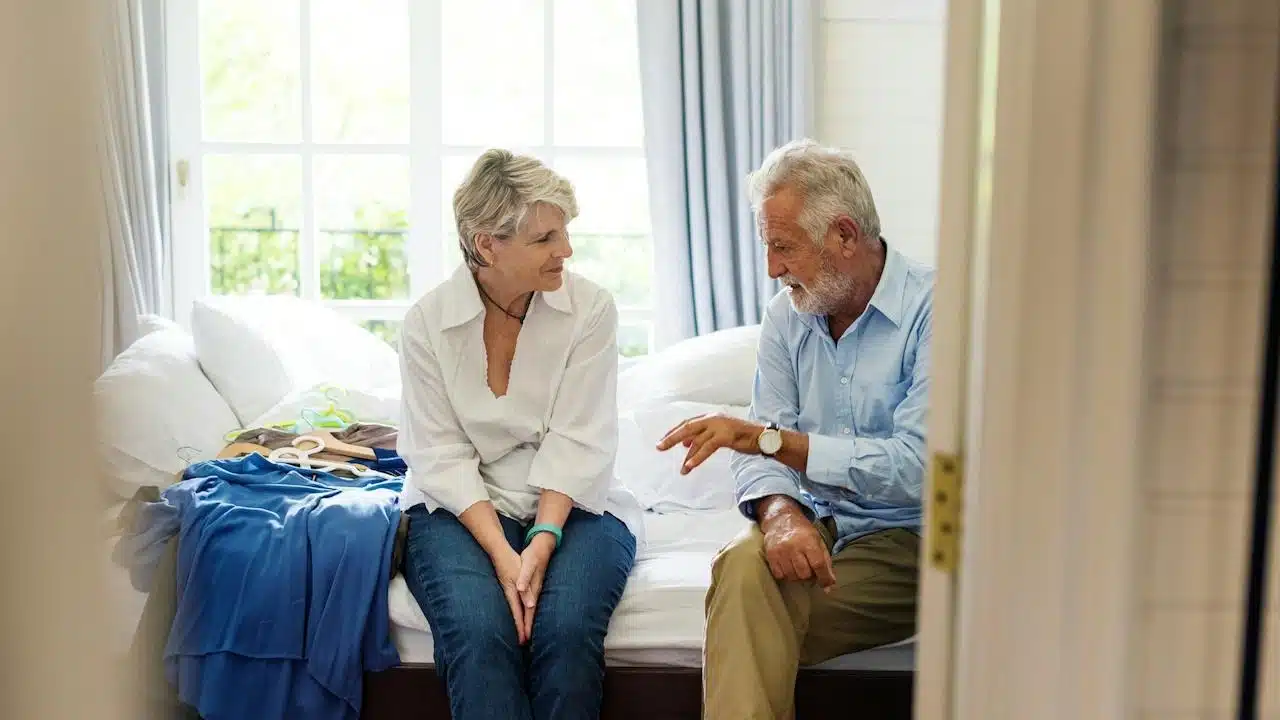 An older couple sitting on a bed and having a conversation.