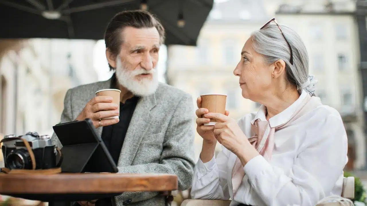 A senior couple sits at an outdoor cafe table, drinking coffee and talking.