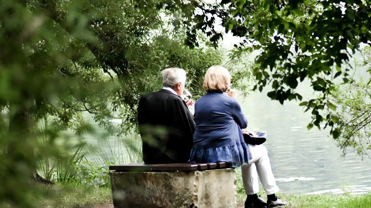 A couple sitting by a lake on a bench and having a drink.