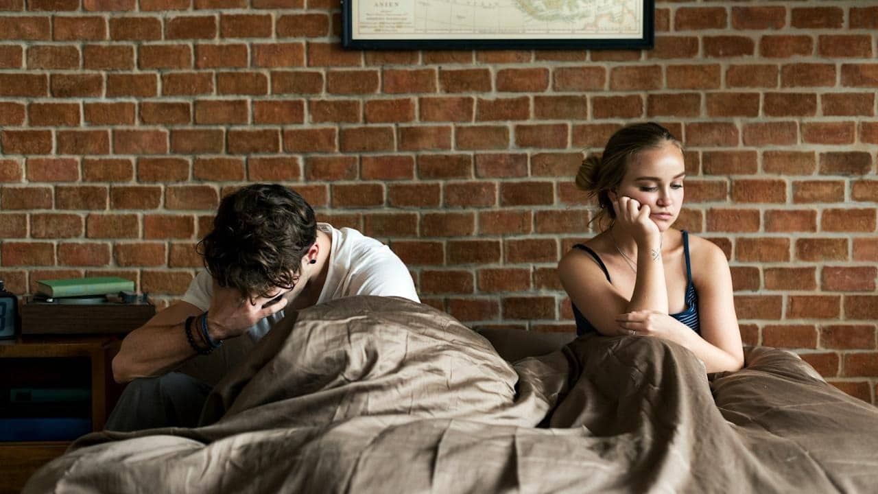A sad couple sitting on a bed after having an argument.