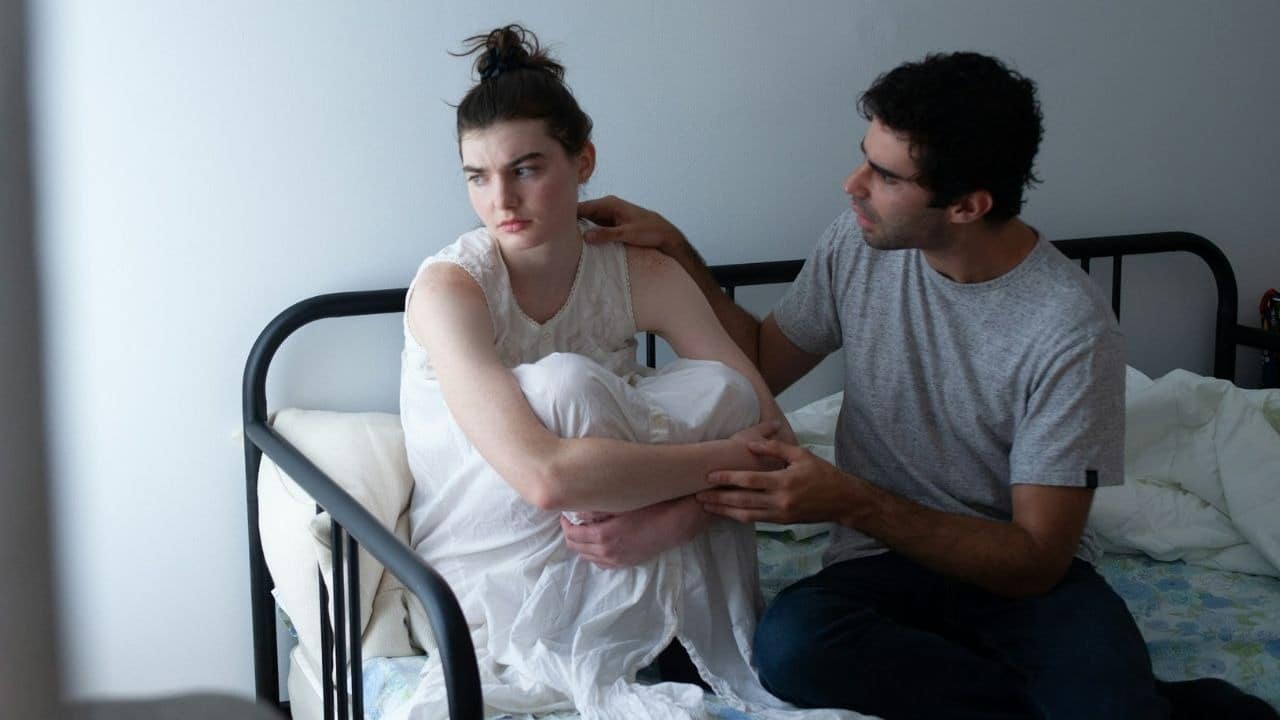 A woman looking away from a man who is trying to comfort her on a bed.