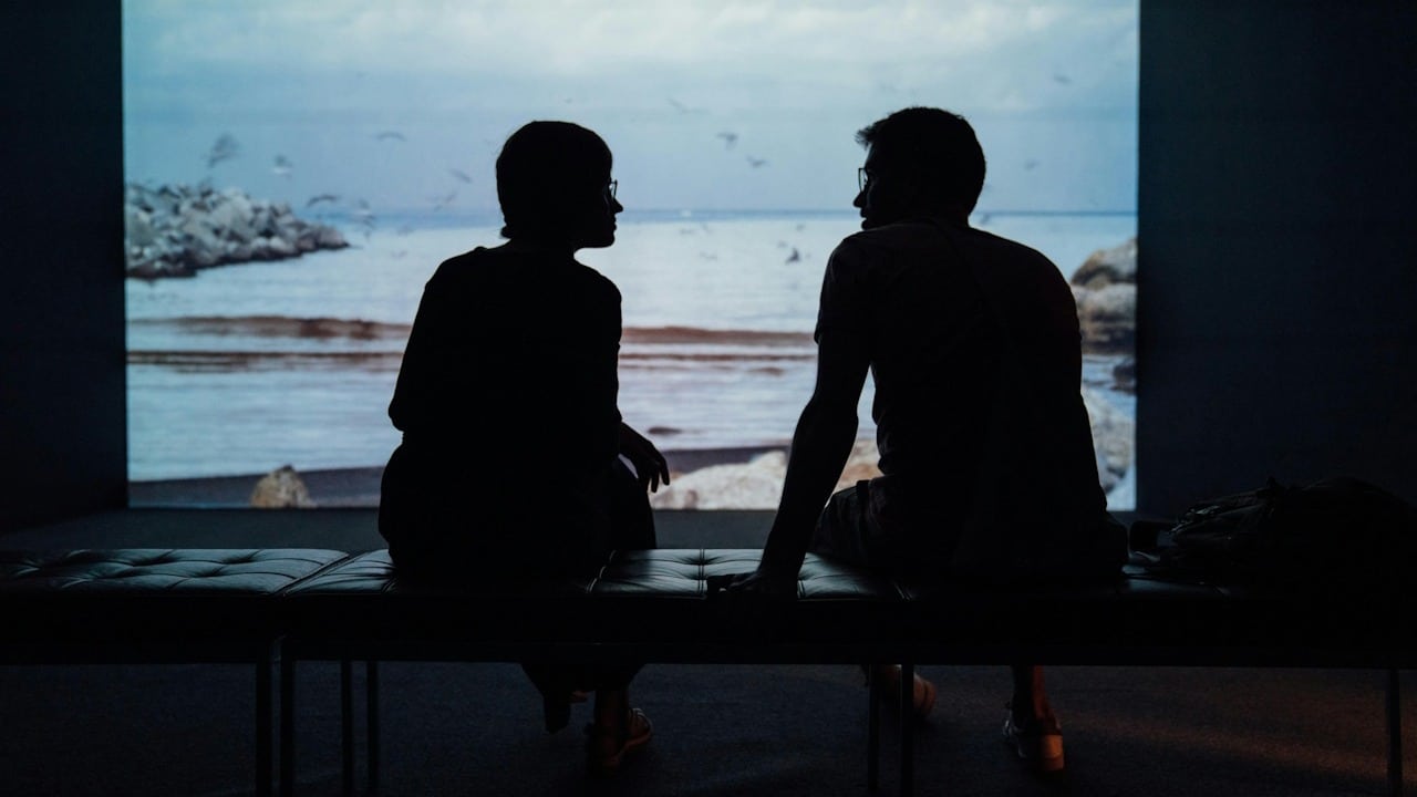Silhouettes of a couple sitting on a bench, facing each other against a projection screen showing a beach scene.