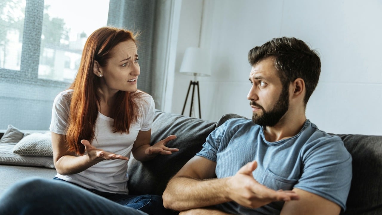 Couple arguing on a couch, both gesturing with their hands during a heated discussion.